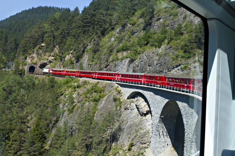 Een Zwitserse gletsjerexprestrein rijdt over een hoog viaduct een tunnel in, gezien vanuit de trein
